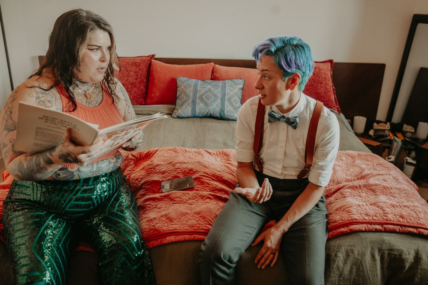 A couple sits together on a bed during a casual indoor photoshoot with natural lighting.