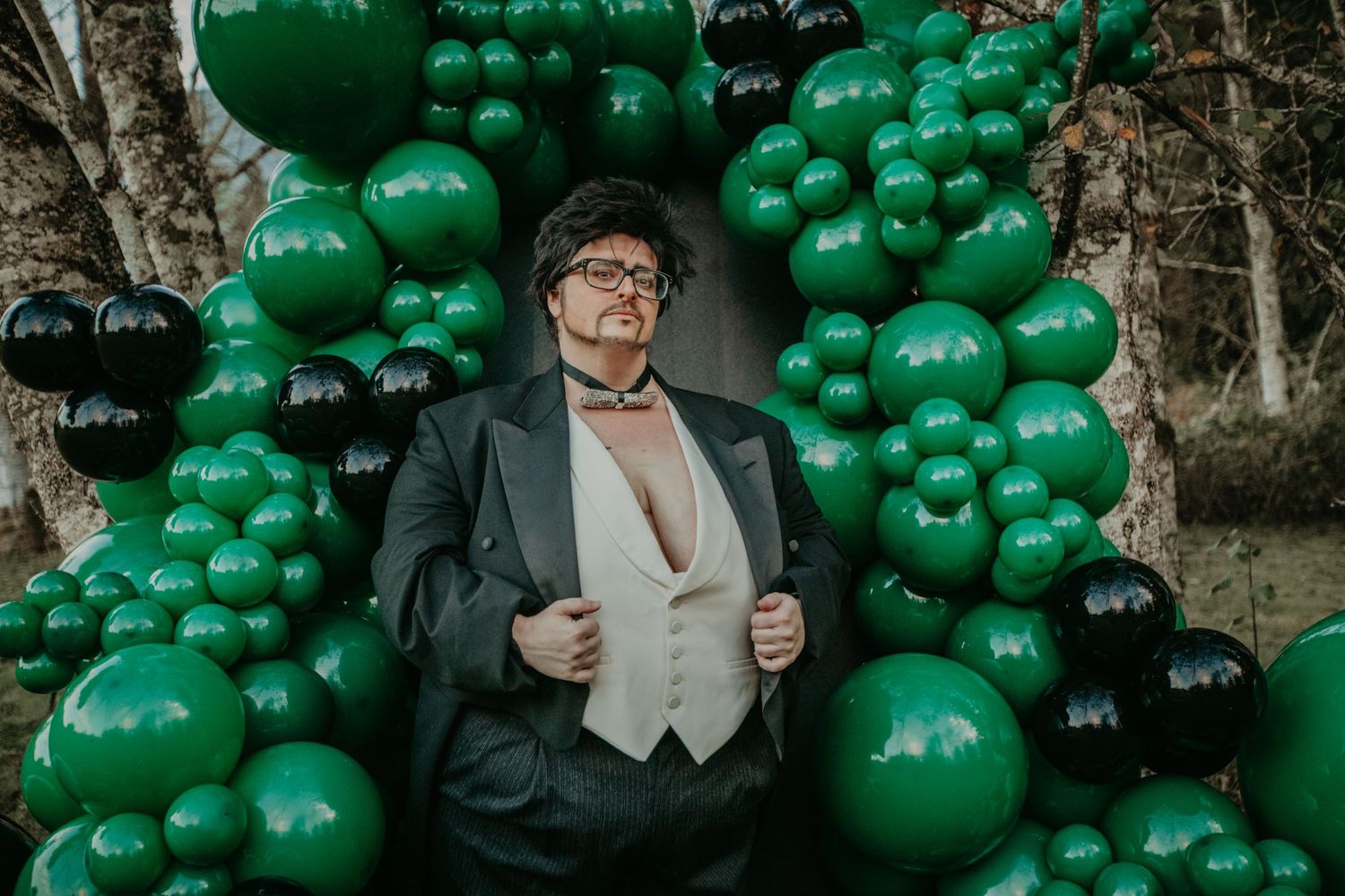 A figure stands against a backdrop of emerald green balloons and birch trees wearing formal evening attire.