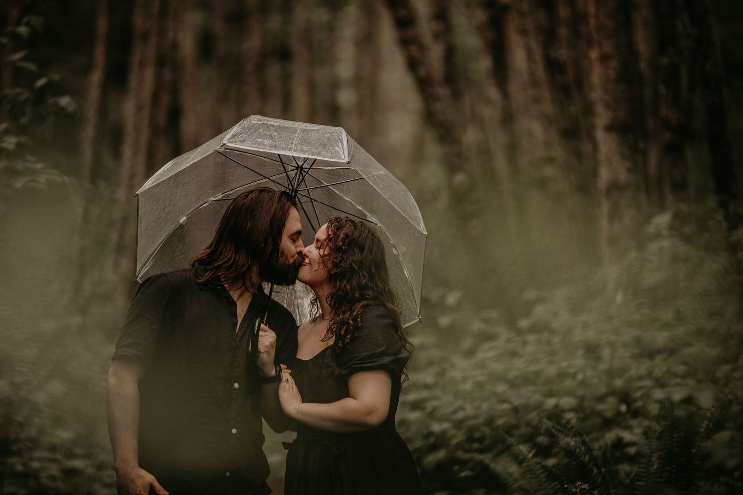 A romantic couple shares an intimate moment under a clear umbrella in a misty forest setting.