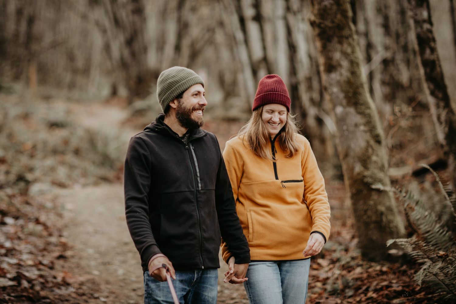 A couple in warm fall attire walking together on a wooded autumn path while sharing a laugh.