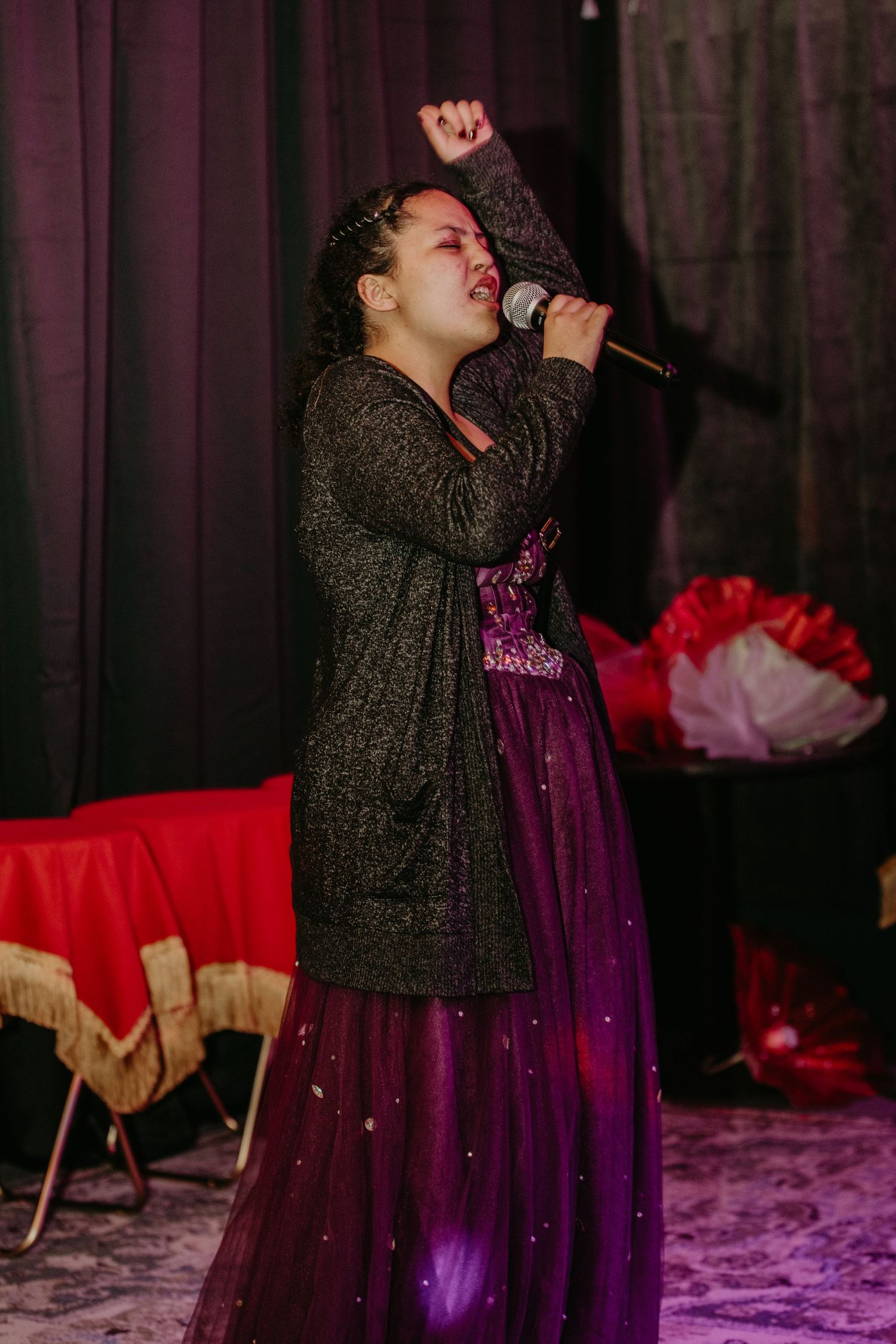 A performer in a purple sparkly dress and dark cardigan sings passionately on stage with a microphone against dark curtains.