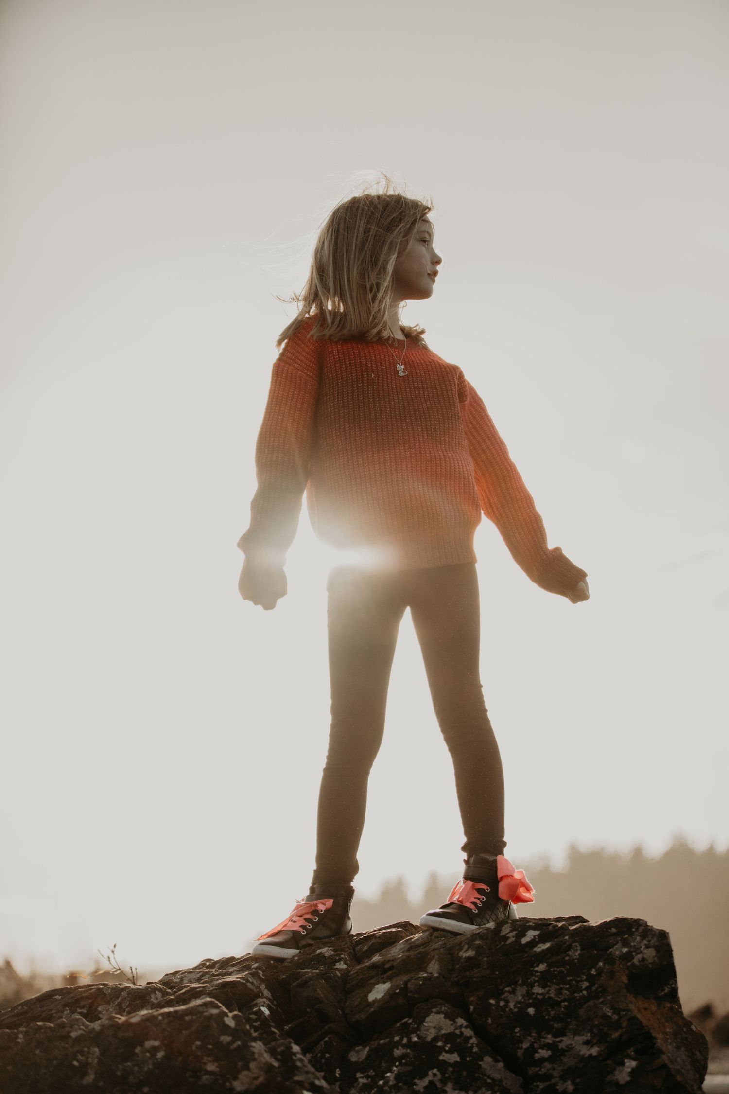 Silhouetted figure in orange sweater stands triumphantly on rocky outcrop during golden hour with sunlight behind them.