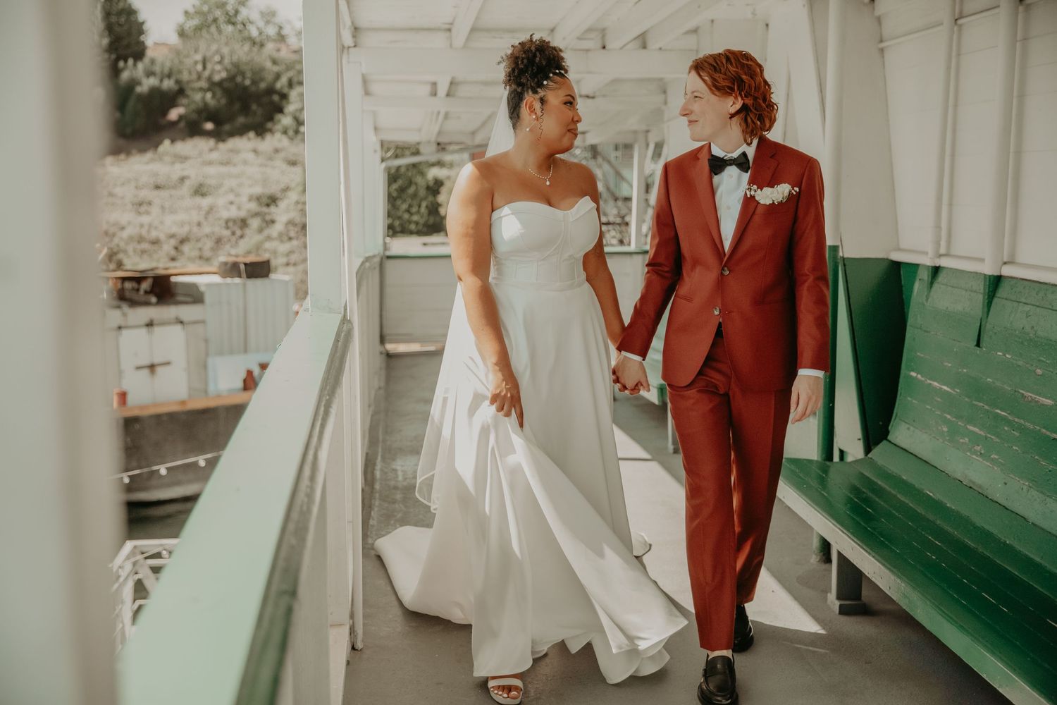 A couple in wedding attire walks together on a covered porch with white pillars and green benches.