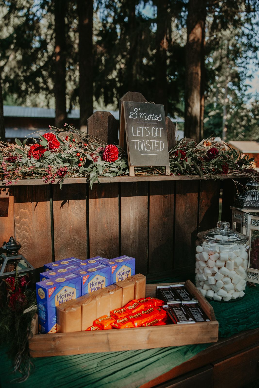 Rustic wooden snack bar display with s'mores ingredients and red floral decorations at an outdoor event.