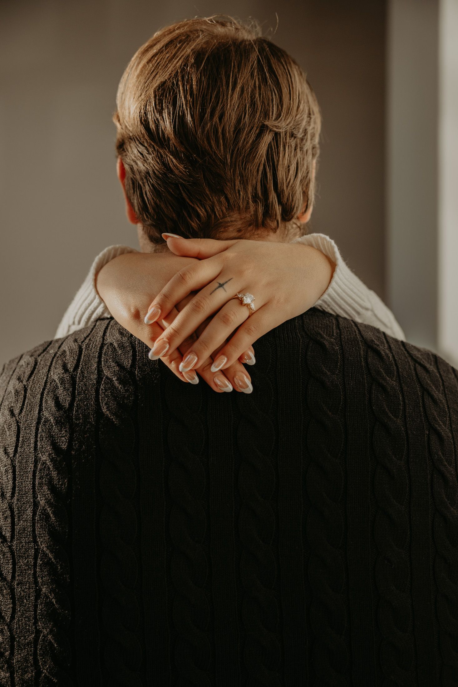 A hand with an engagement ring rests on the neck of a person wearing a dark cable-knit sweater, viewed from behind.