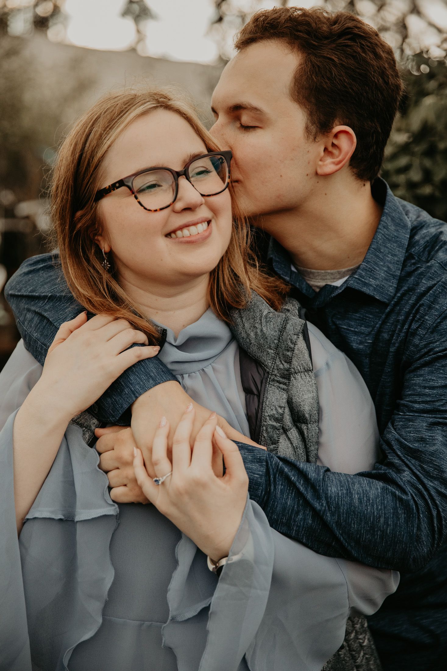 A young couple shares a tender moment outdoors, man kissing woman's cheek as she smiles wearing glasses and blue dress.
