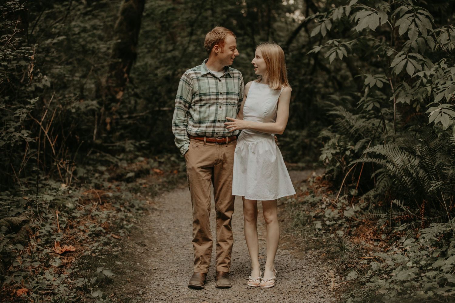 Romantic couple walking together on an autumn forest path lined with lush greenery and fallen leaves.