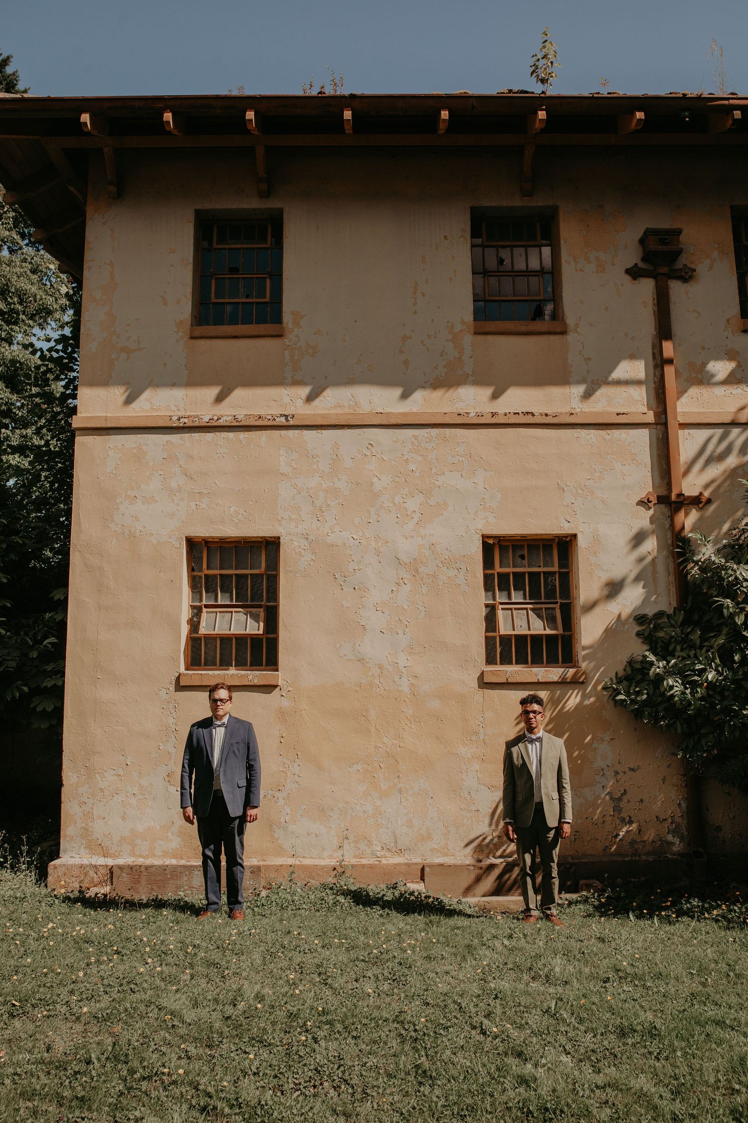 Two figures stand at opposite ends of a beige building wall against a green grassy hillside.
