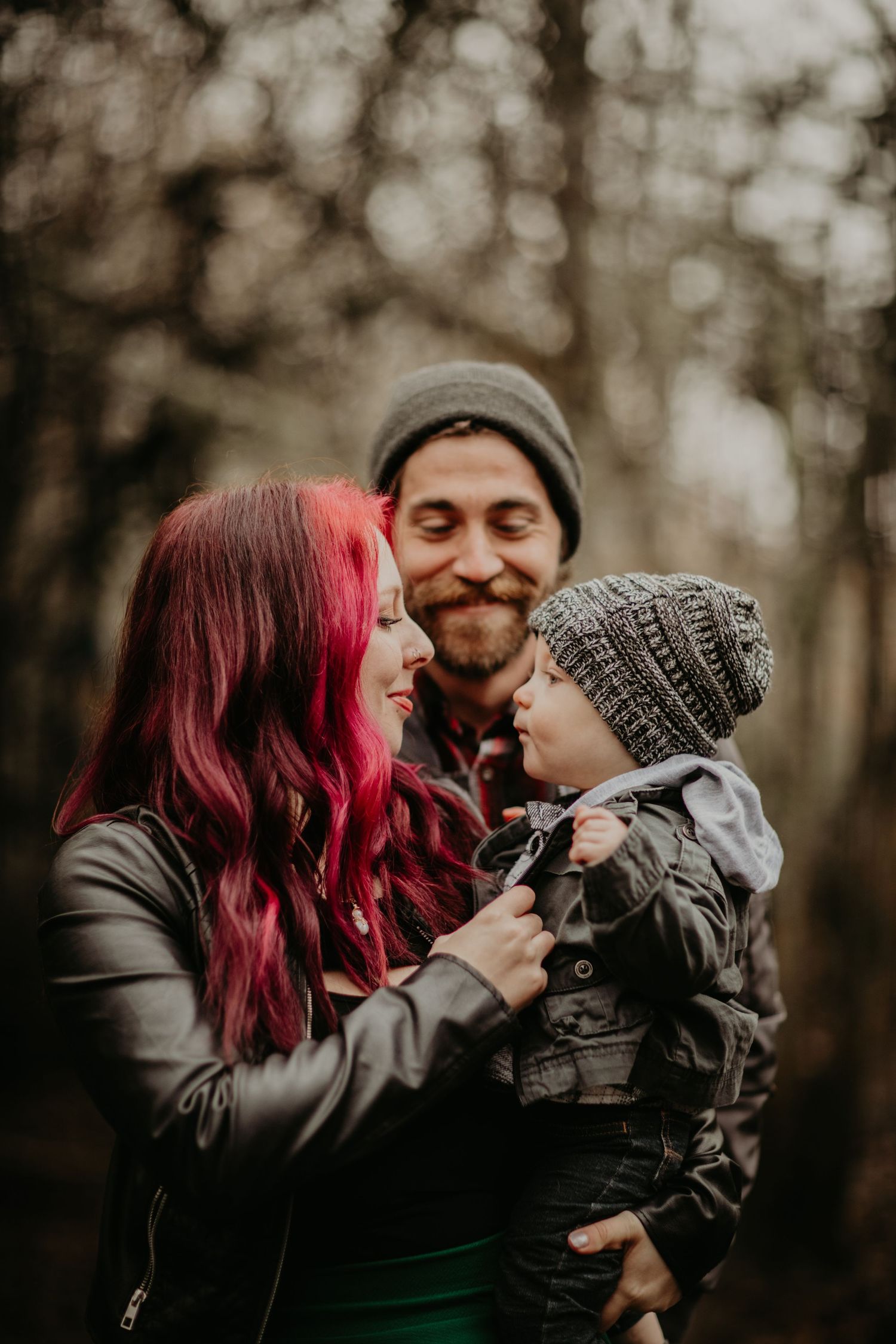 A family shares a tender outdoor moment in fall wearing knit beanies among blurred trees in the background.