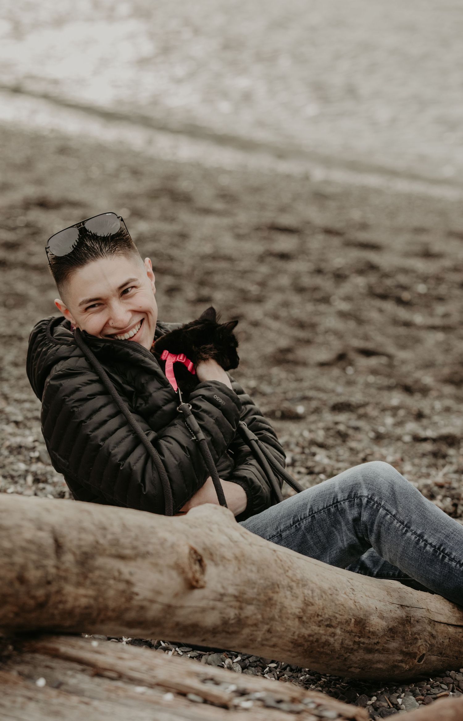 A person sits on a beach log wearing a black jacket while holding a black cat with a pink collar.