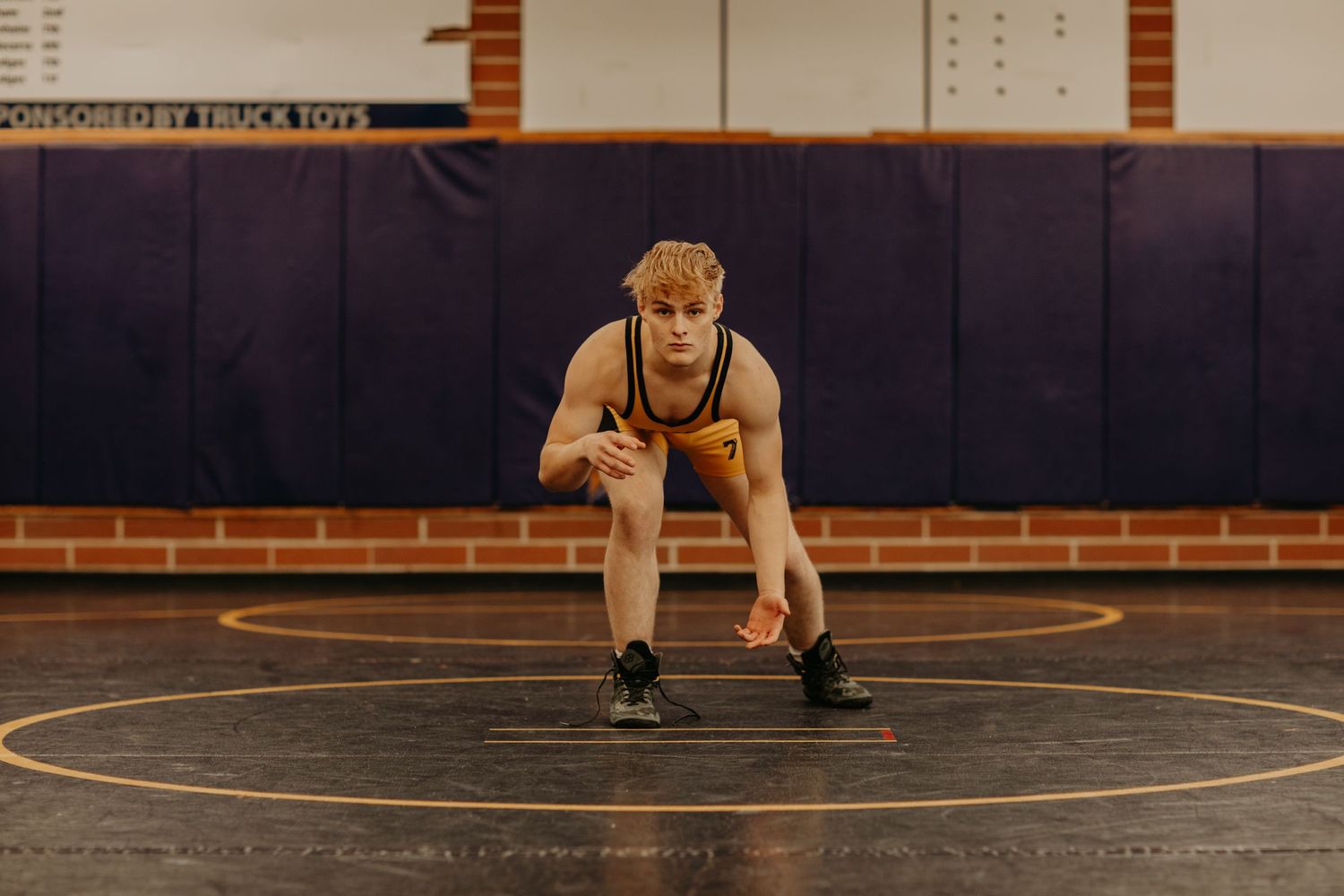 Wrestler in tan singlet crouches in ready position on wrestling mat in dimly lit gymnasium with brick wall background.