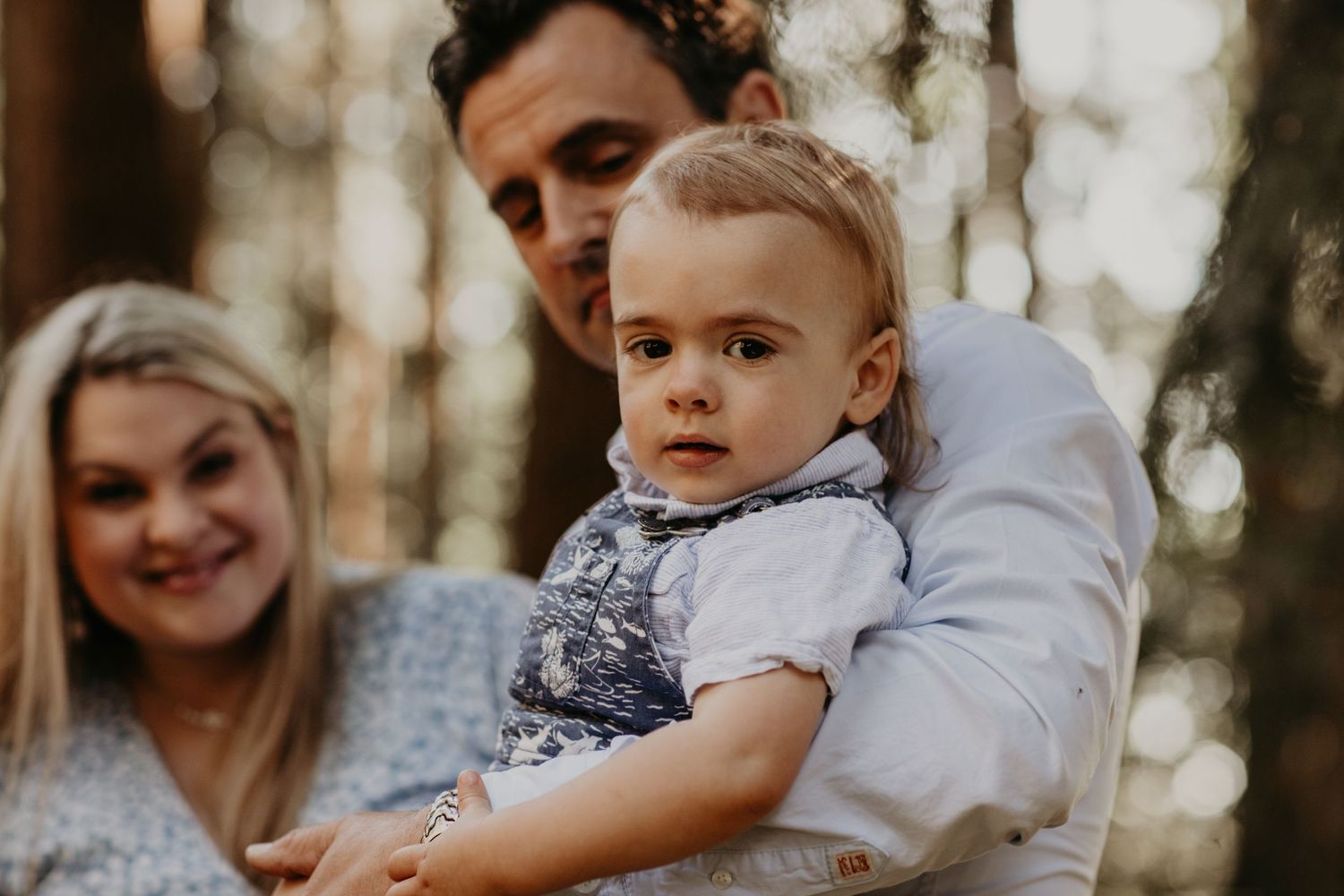 A toddler in a gray t-shirt held up outdoors with a soft-focused forest background.