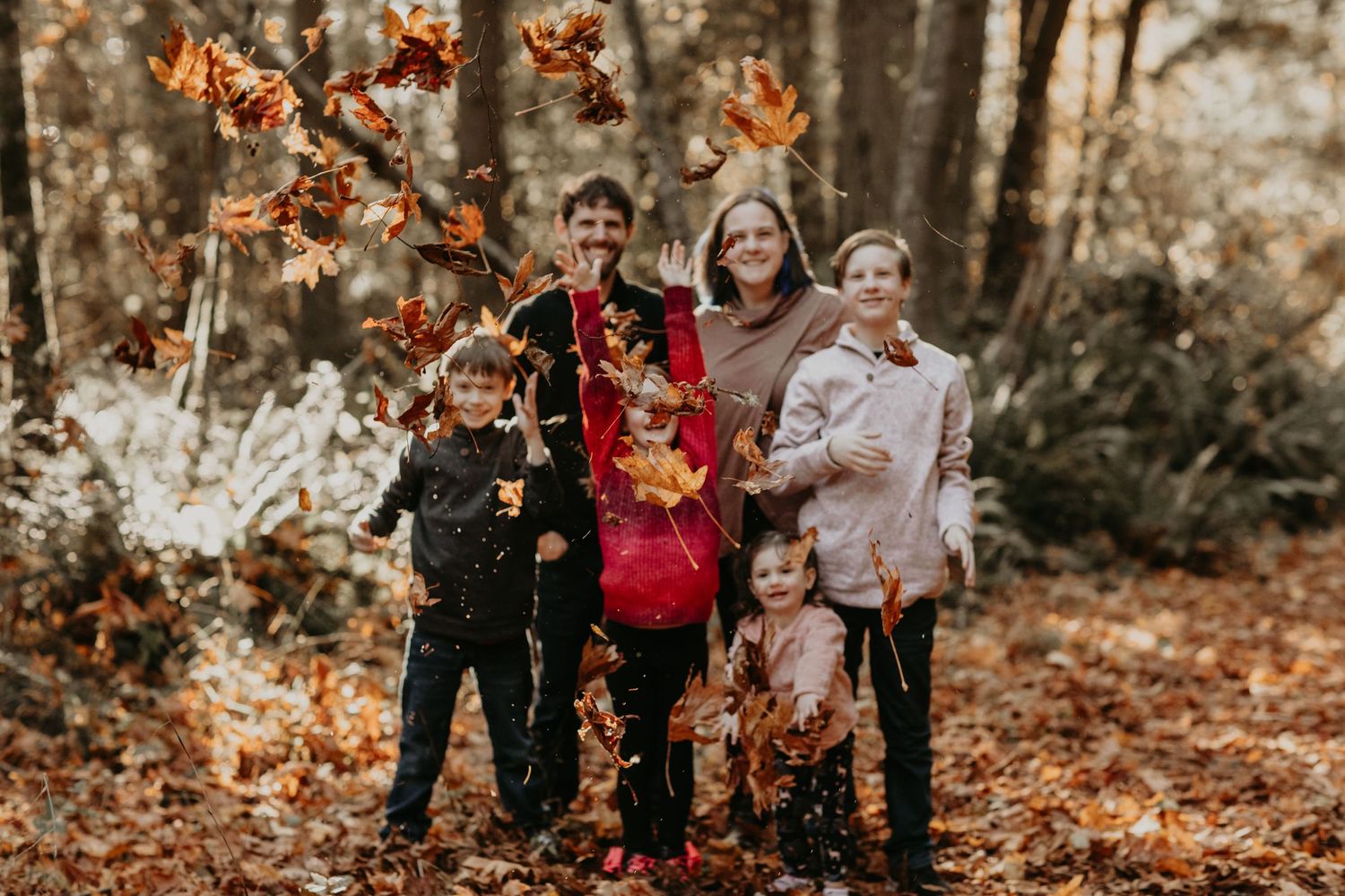A group stands together in warm autumn clothing against a backdrop of falling leaves in a wooded area.