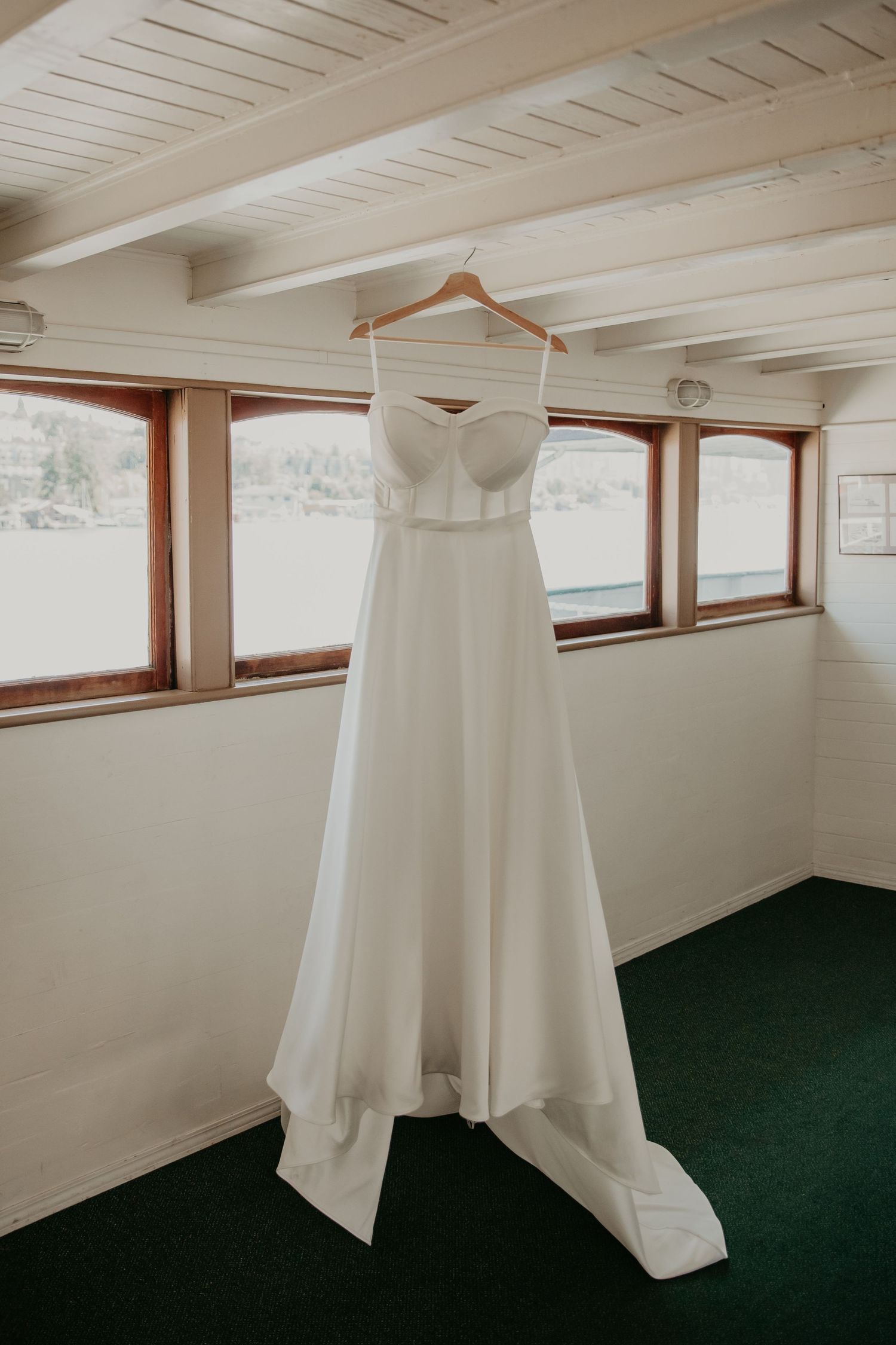 White wedding dress hanging in a room with wooden beams and green carpeting.