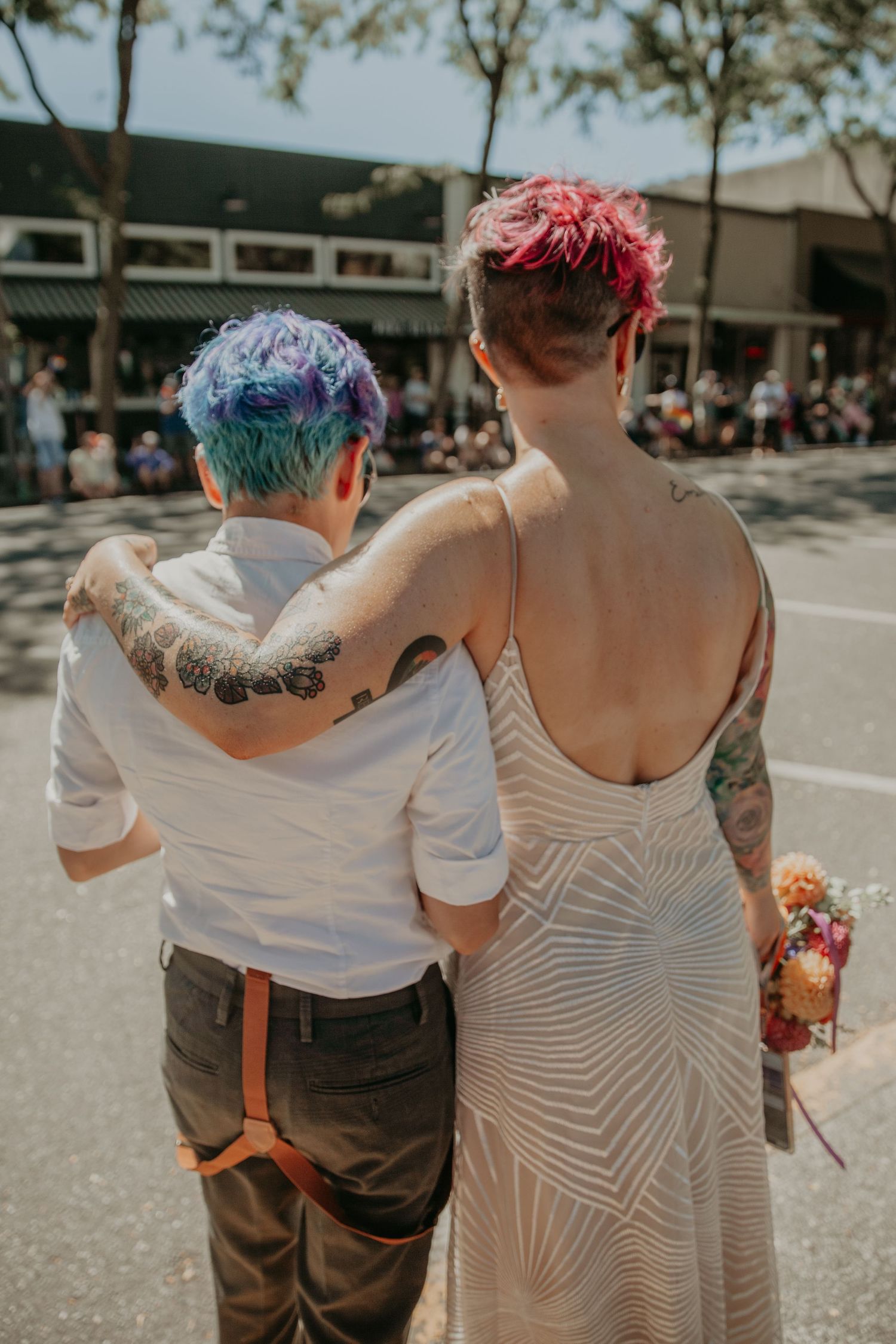 A couple with colorful hair and tattoos walking together in casual attire on a sunny urban street.