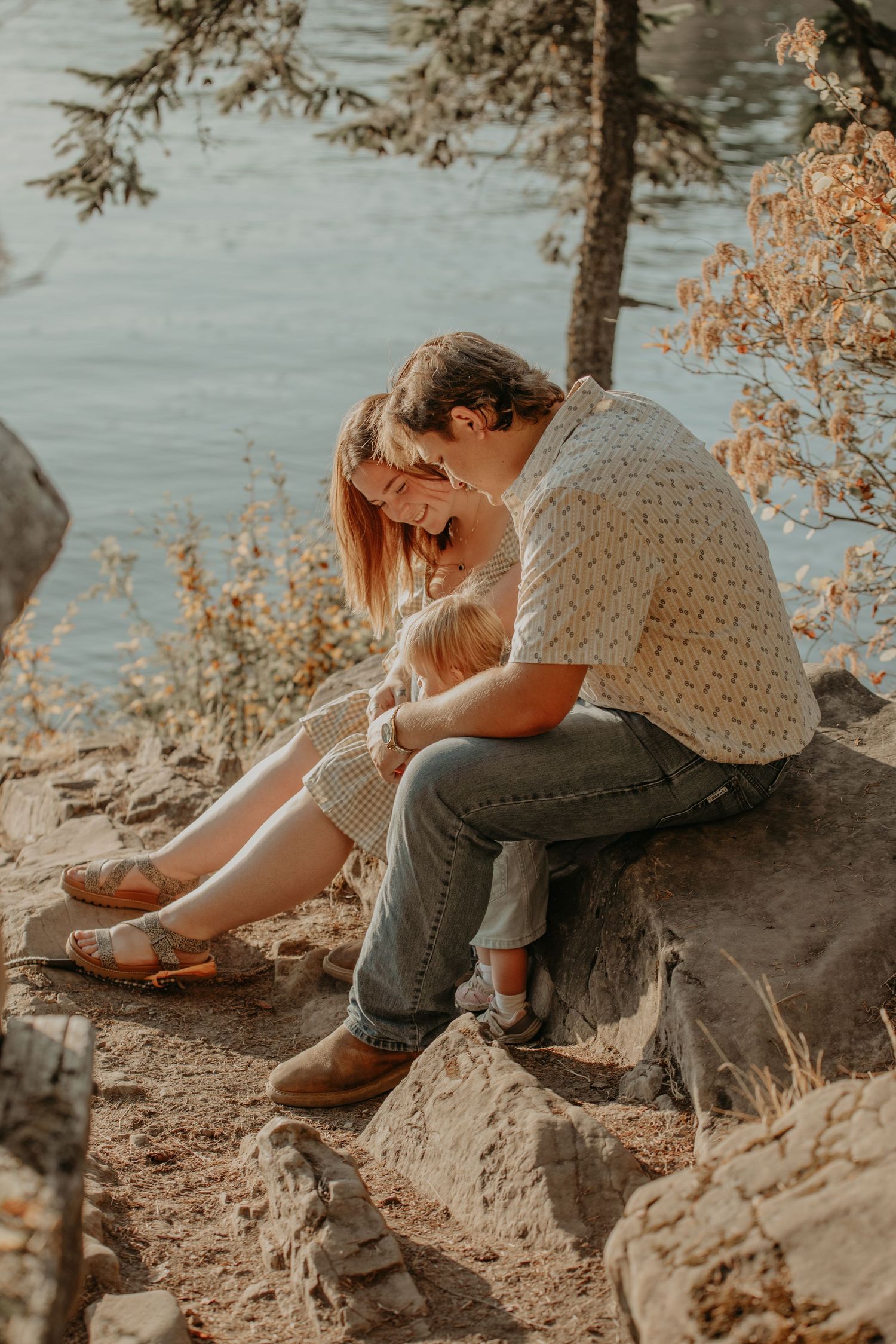 Moody autumn sequence showing person sitting on riverside rocky slope wearing knit sweater and jeans against water backdrop.
