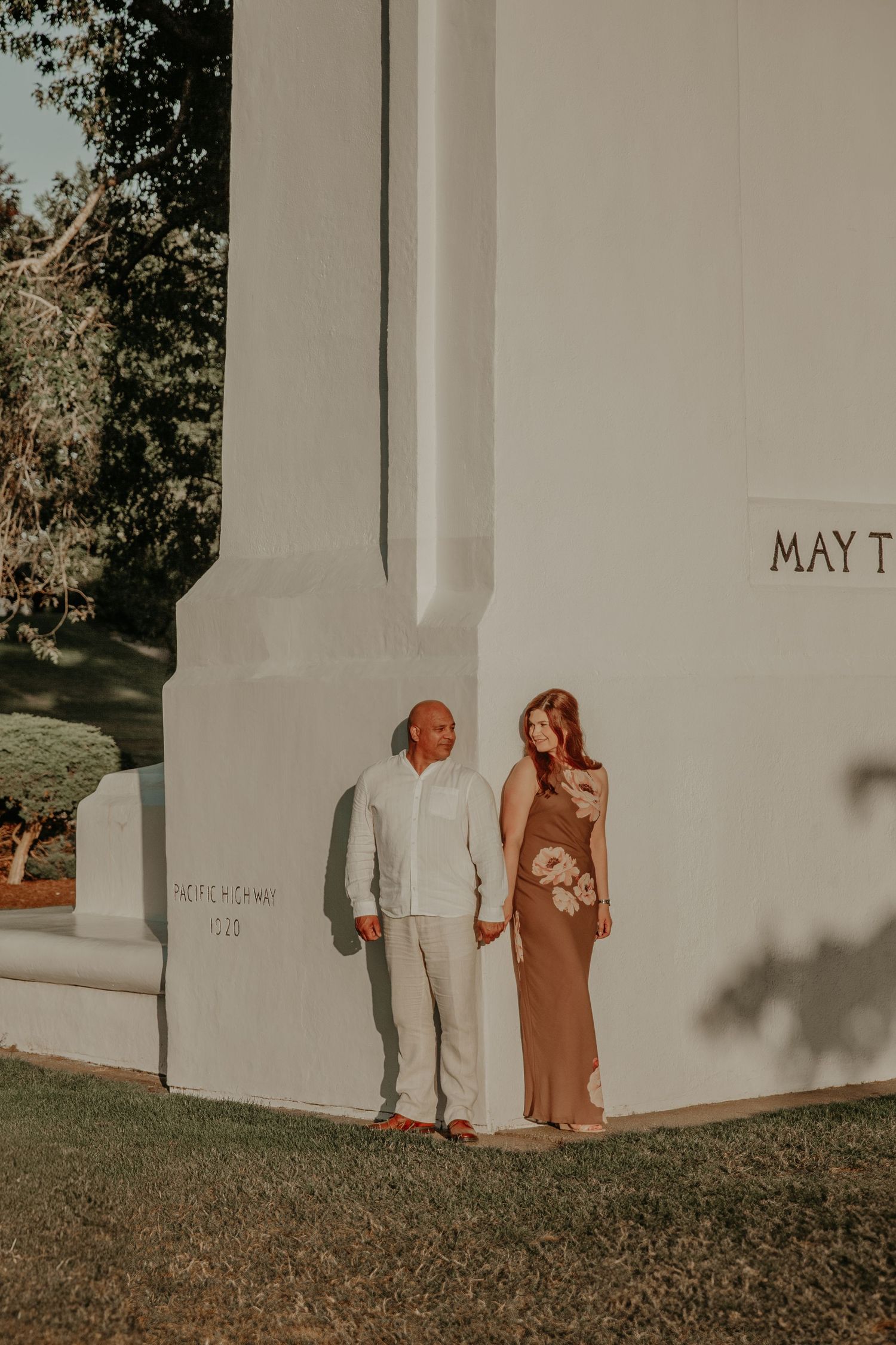 A romantic couple standing by a white church exterior wall bathed in golden sunlight and shadow play.
