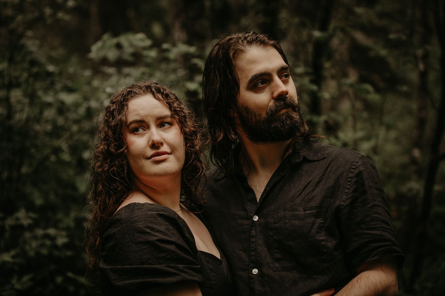 An intimate portrait photograph of a couple with curly hair against a moody forest backdrop in dark attire.