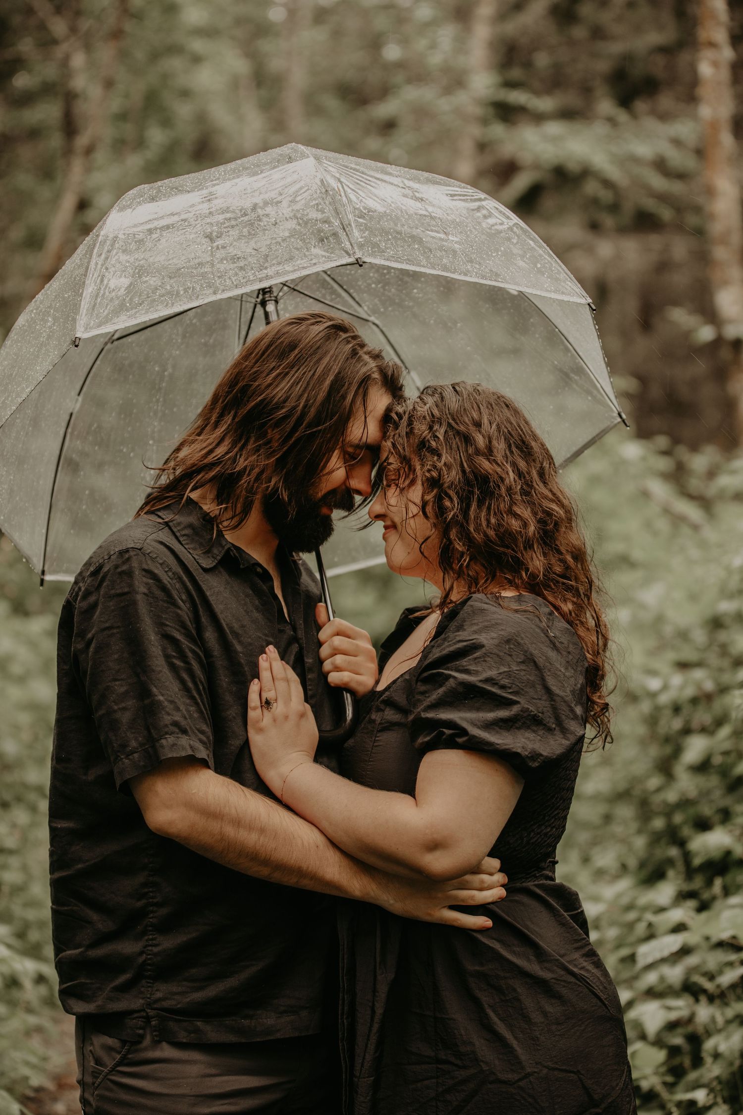 Romantic couple embracing under clear umbrella in forest setting with moody lighting.