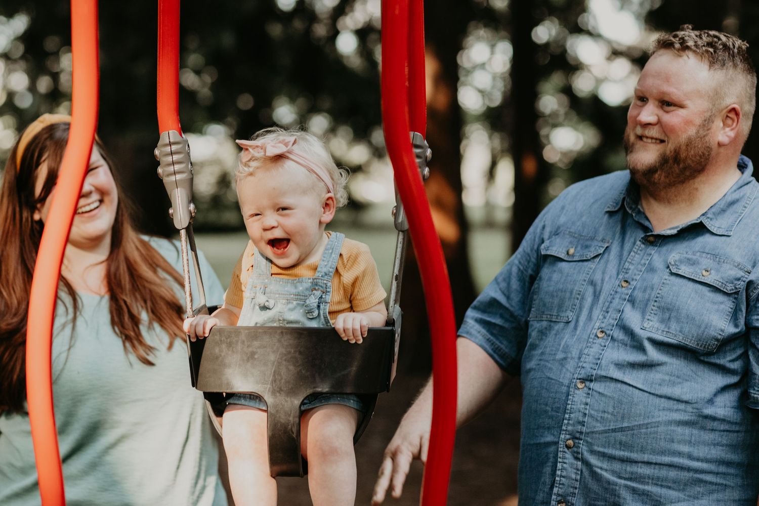 Happy family having fun together with a toddler on a red swing in a park on a sunny day.