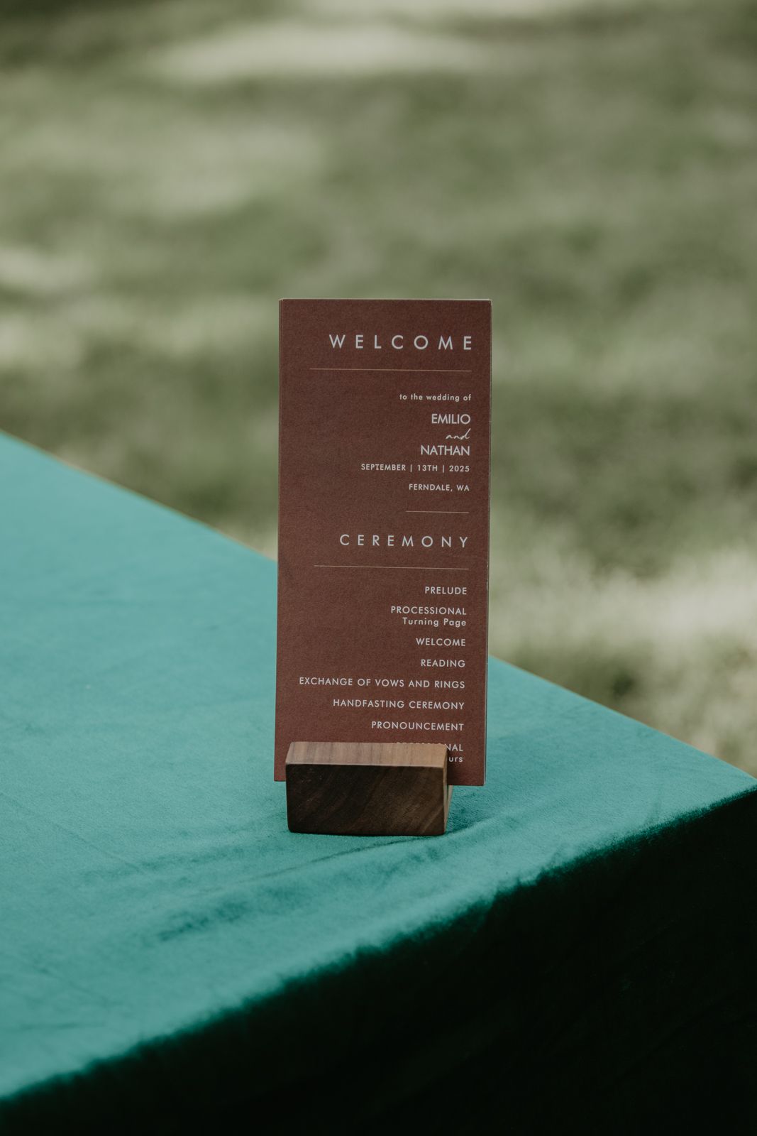 A brown wedding ceremony program card stands upright on a wooden holder against a turquoise surface with blurred background.
