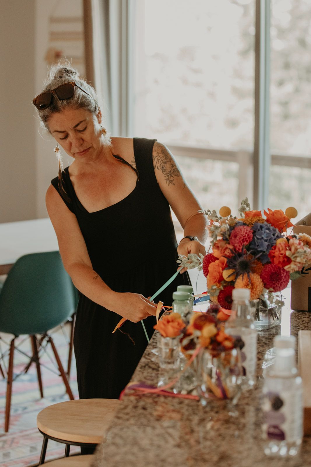 Florist arranging vibrant red, orange and purple flowers in glass vases on a table near a bright window.