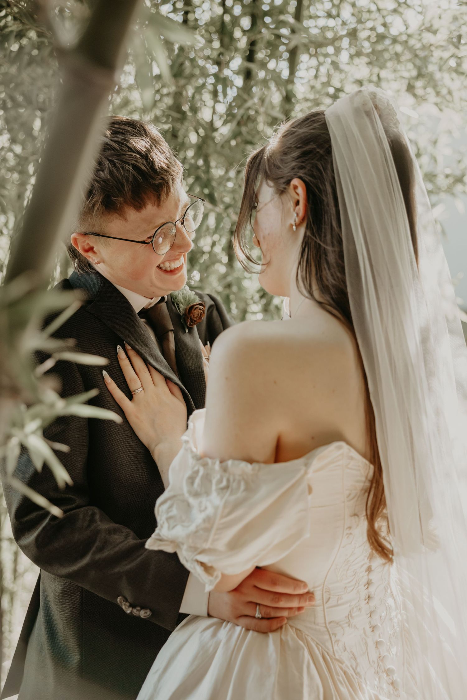 Bride in off-shoulder gown and veil smiles with groom in dark suit among green trees.