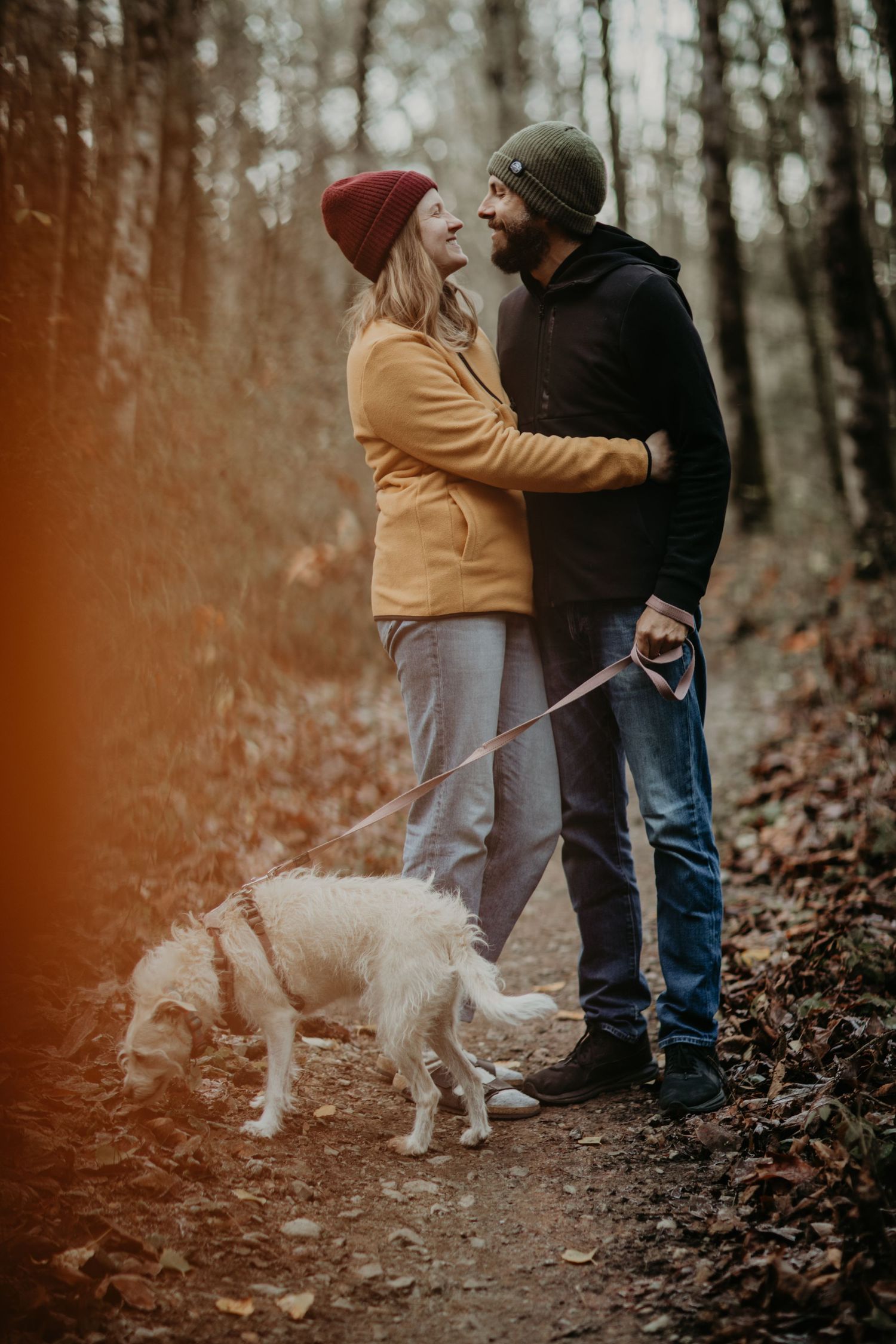 A couple in winter coats walking a white dog on an autumn forest trail with warm lens flare.