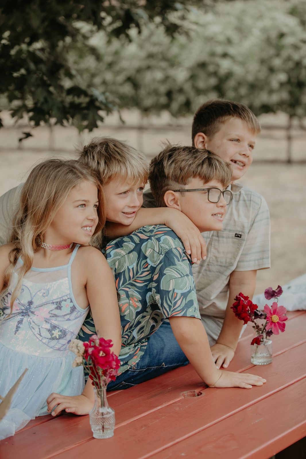 Children lined up at a red picnic table with pink flowers in mason jar vases on a sunny summer day.
