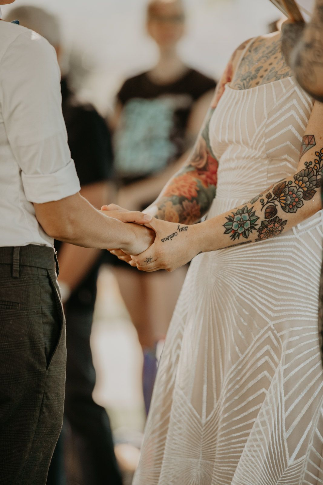 Close-up of hands holding during a wedding ceremony, with one person wearing a geometric white dress with tattoos.