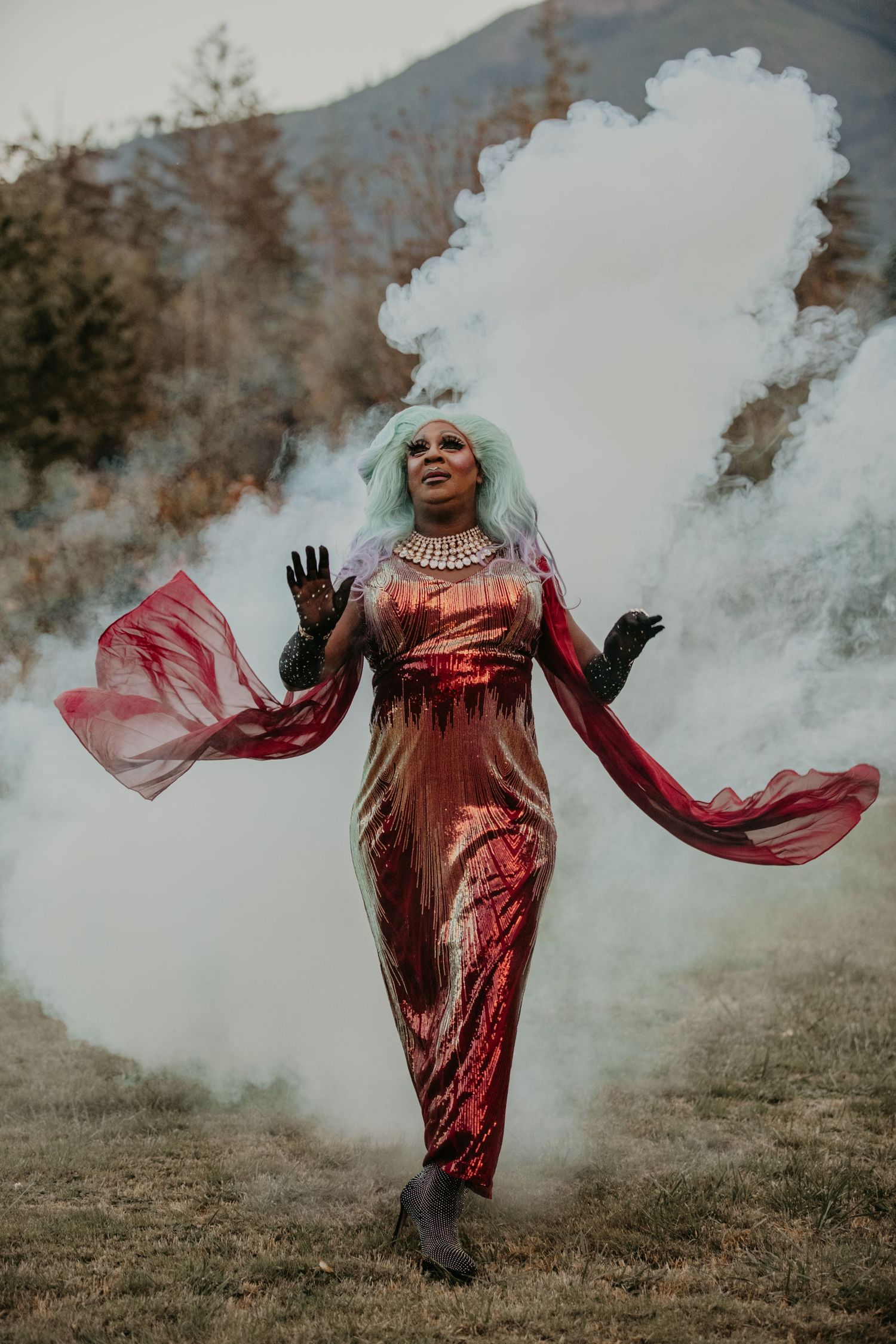 Artistic fashion photo with flowing red dress and white smoke effects in mountain landscape.