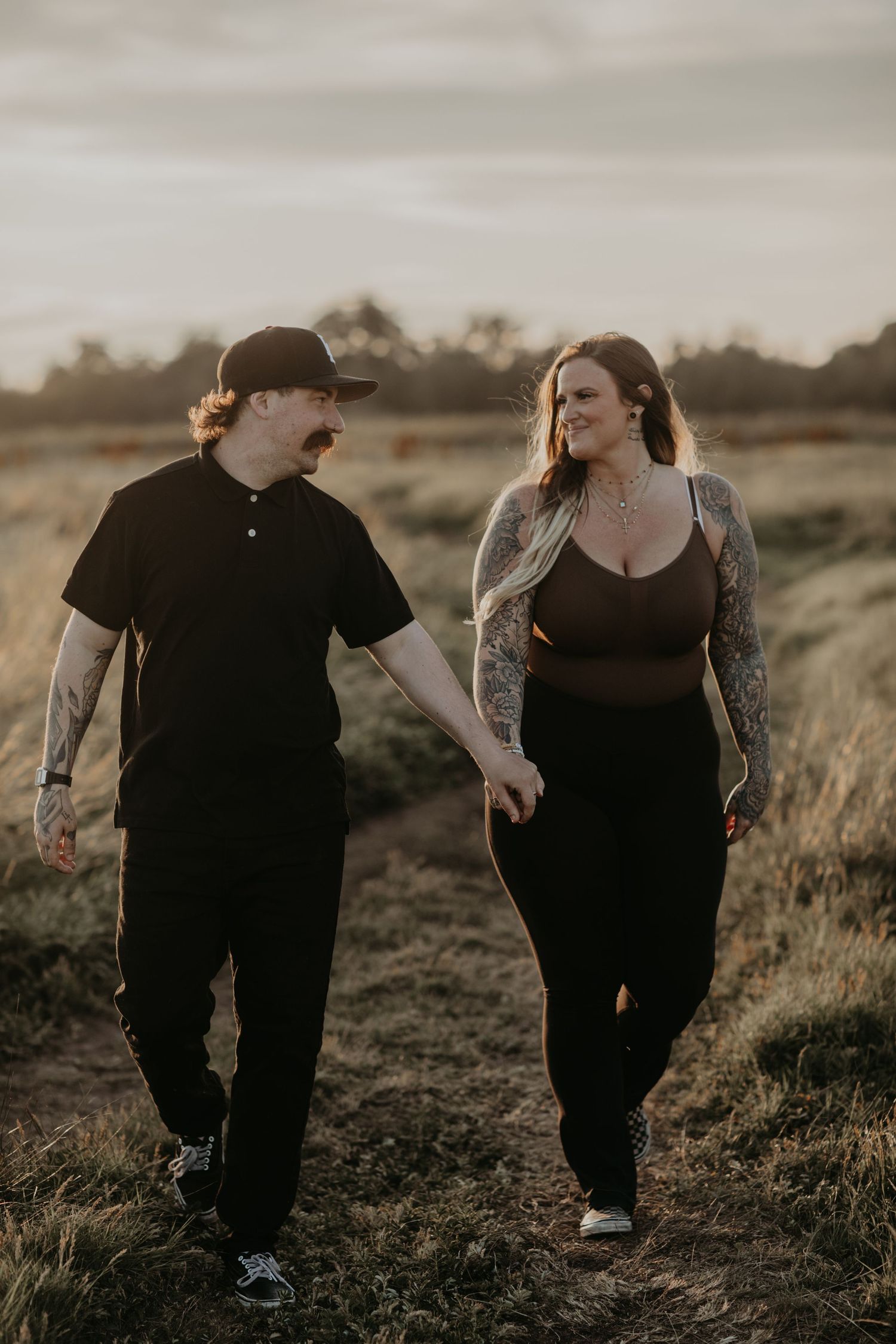 Couple walking hand in hand through a grassy field at sunset wearing casual black clothing and showing tattoos.