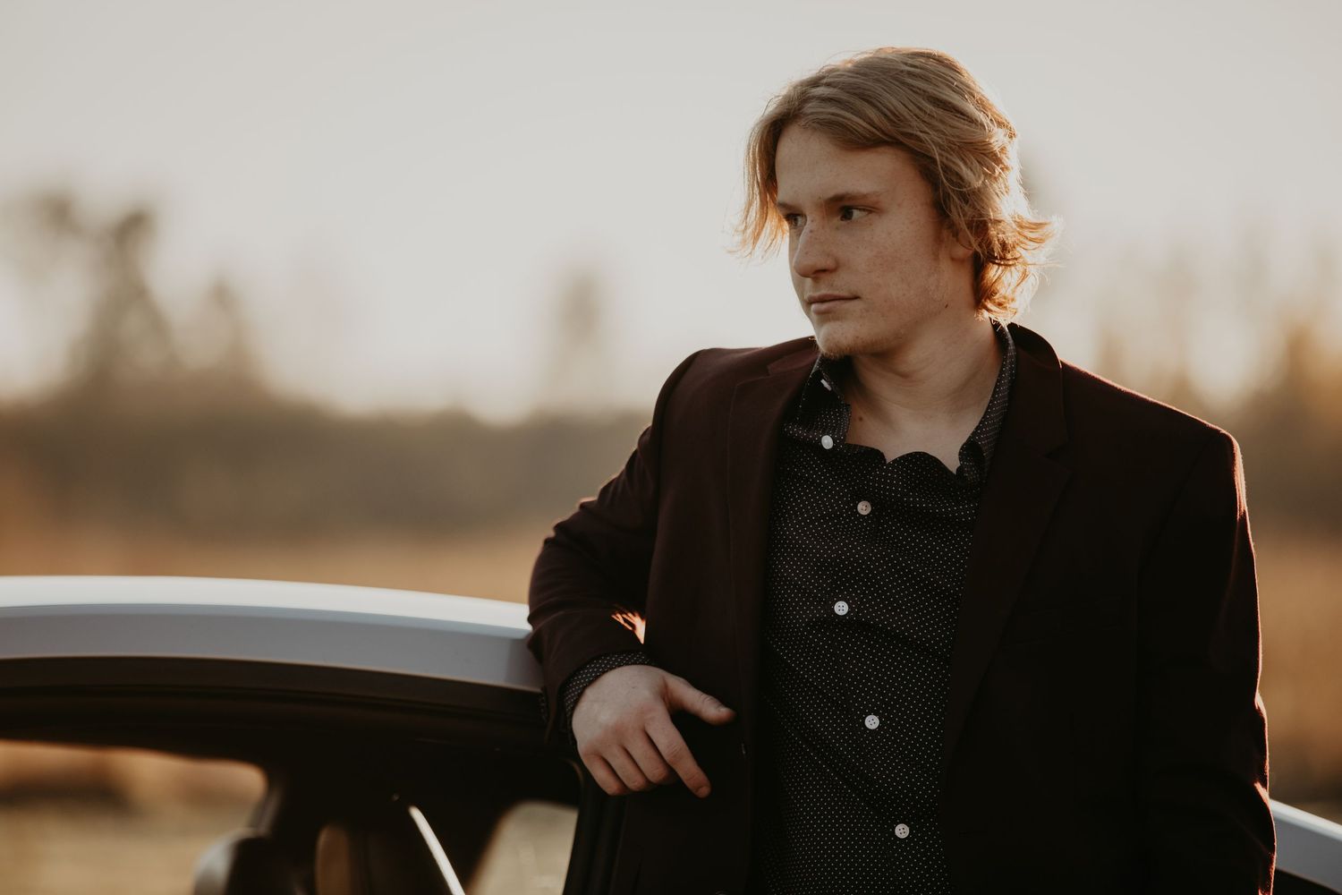 Person wearing a black button-up shirt leans against a car at sunset with warm golden lighting in the background.