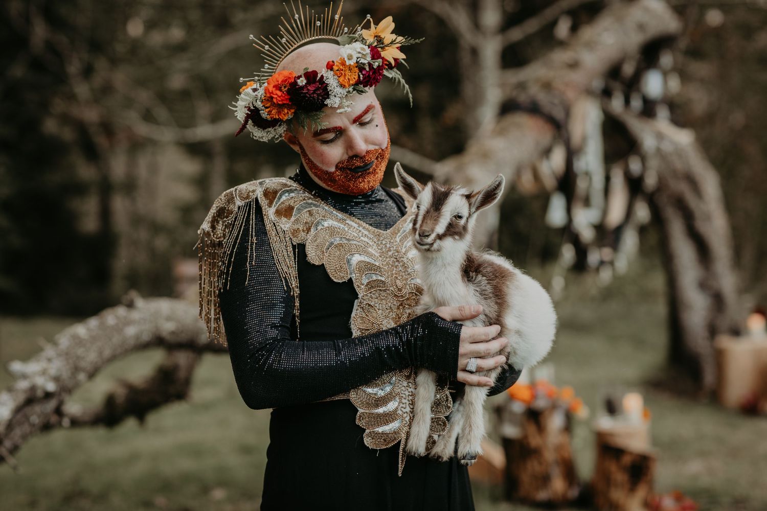 Person wearing whimsical floral headpiece and dark dress holds an adorable gray rabbit in a magical forest setting.