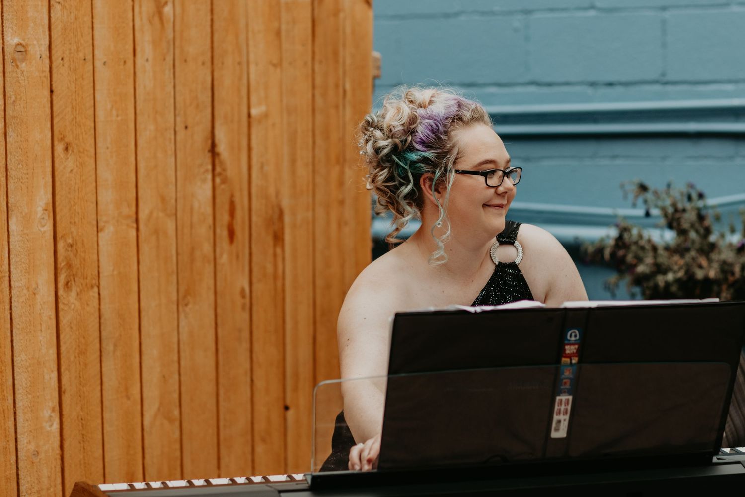 Musician in a one-shoulder black dress plays keyboard against blue wall and wood fence backdrop.