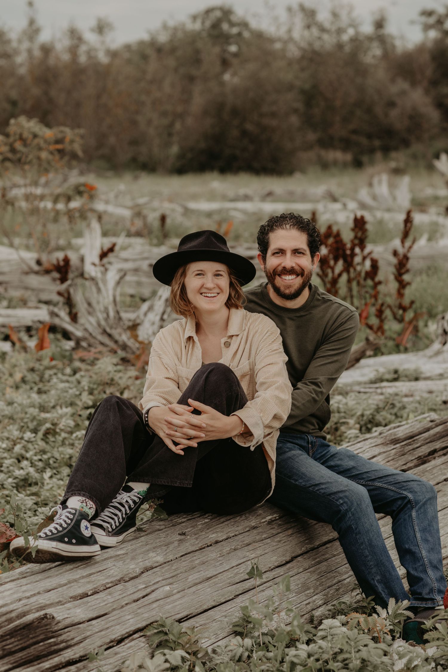 A couple sits together on driftwood at a beach during sunset, dressed casually with her in a black hat and him in green.