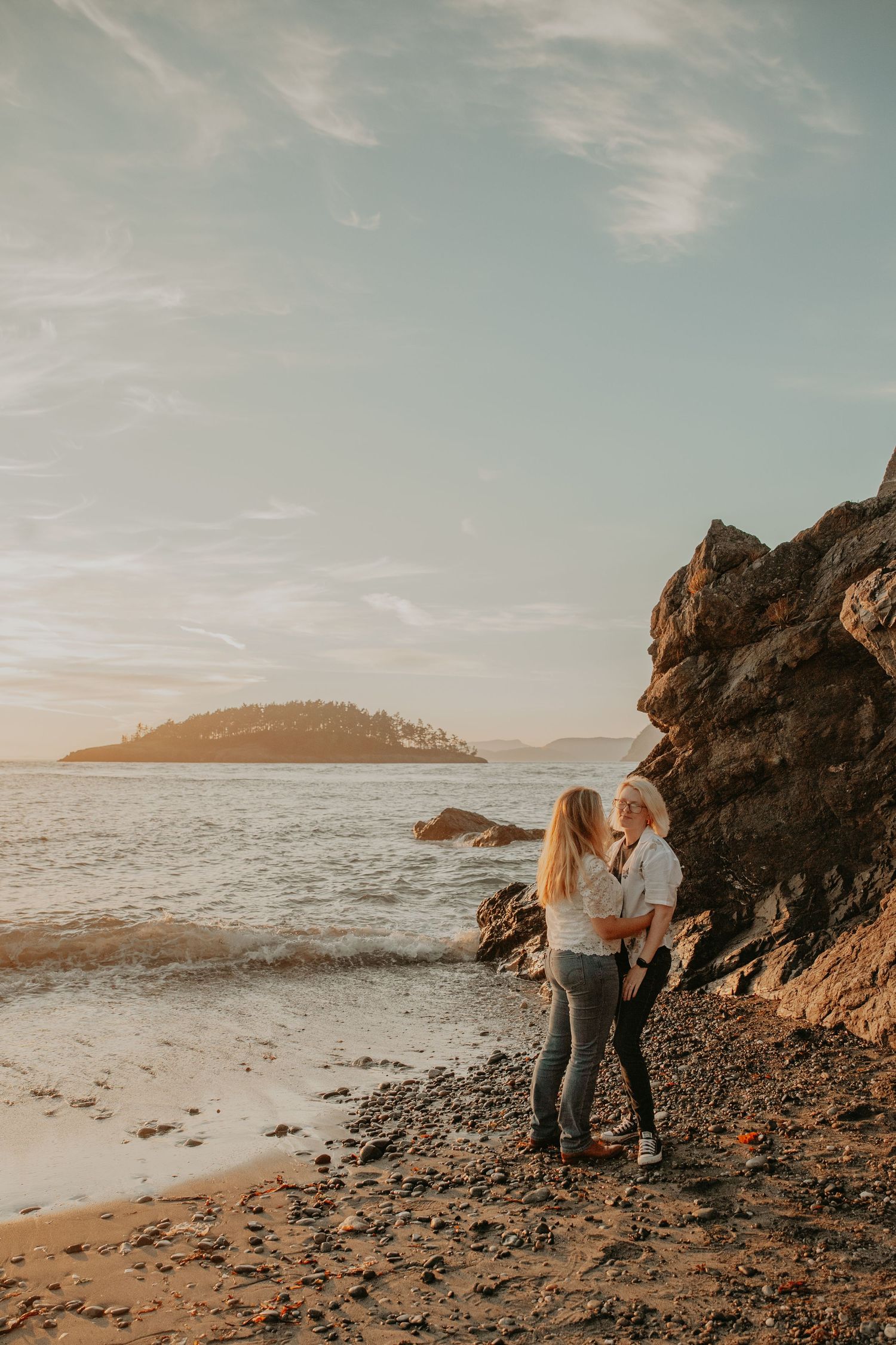 A couple shares an intimate moment at sunset on a rocky beach coastline with dramatic cliffs and ocean views.