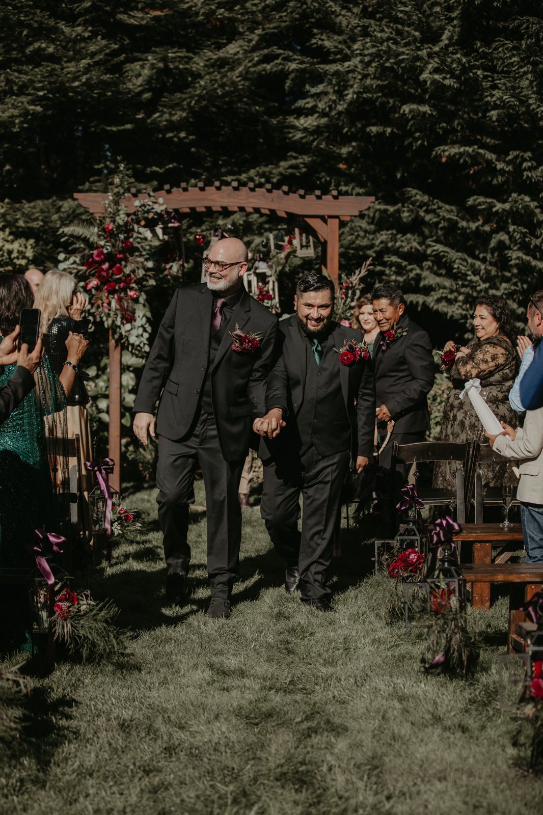 Wedding guests gather on a lawn beneath trees while watching a ceremony under a floral-decorated wooden pergola.