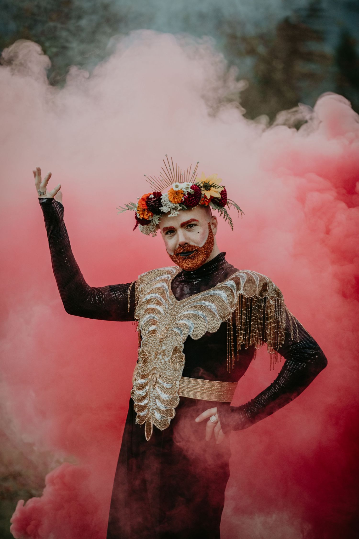 Artistic portrait of a figure in black clothing with white details and floral crown surrounded by pink smoke.