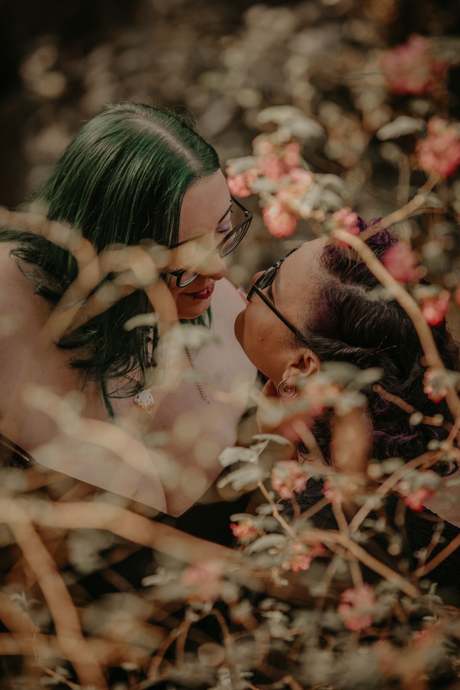Two women with colorful hair and glasses share a romantic moment surrounded by blooming flowers in a moody outdoor setting.