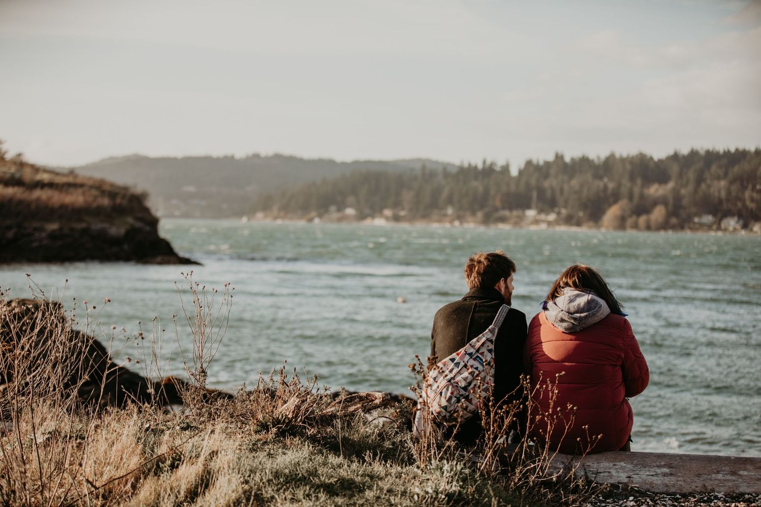 Two people in warm clothing sit together on a cliff overlooking a scenic lake with forested hills in the distance.