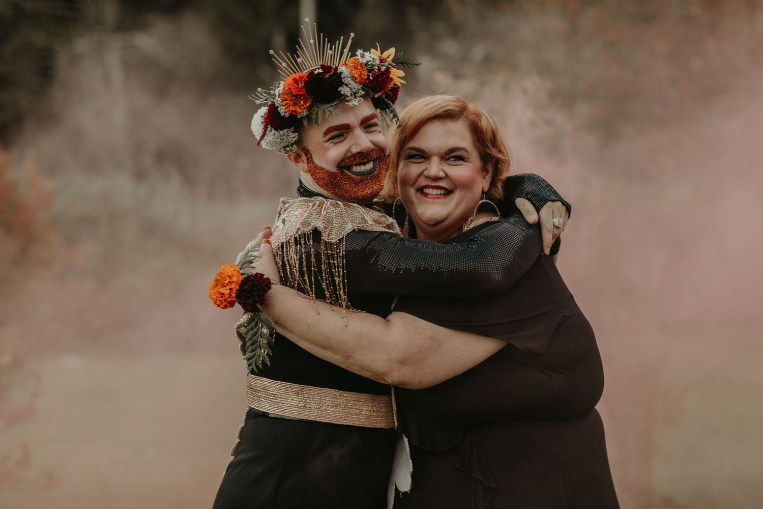Two people embrace joyfully in a moody setting with smoke while one wears flowers in their hair and a dark outfit.