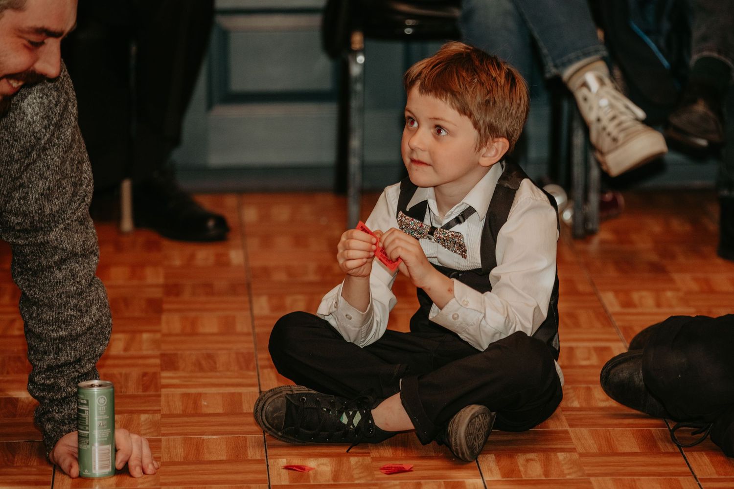 A young child in formal attire sits cross-legged on a hardwood floor during an event.