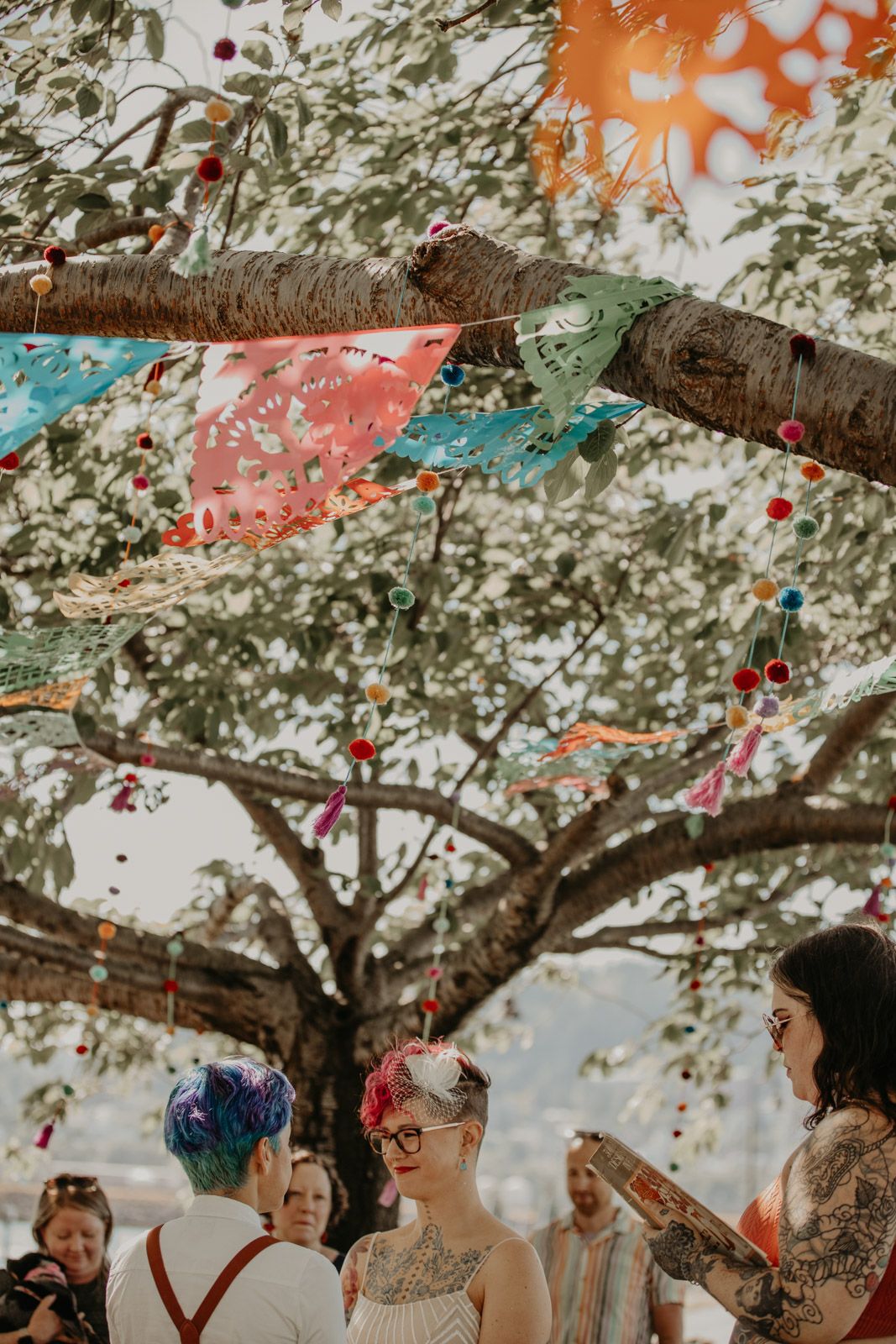 Colorful papel picado banners and decorations hang from tree branches at an outdoor celebration.