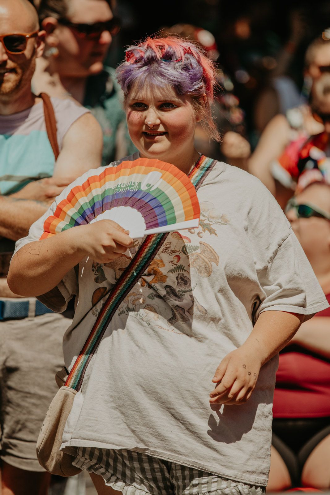 Person in a gray t-shirt holds a colorful rainbow fan while sitting in a crowded audience.