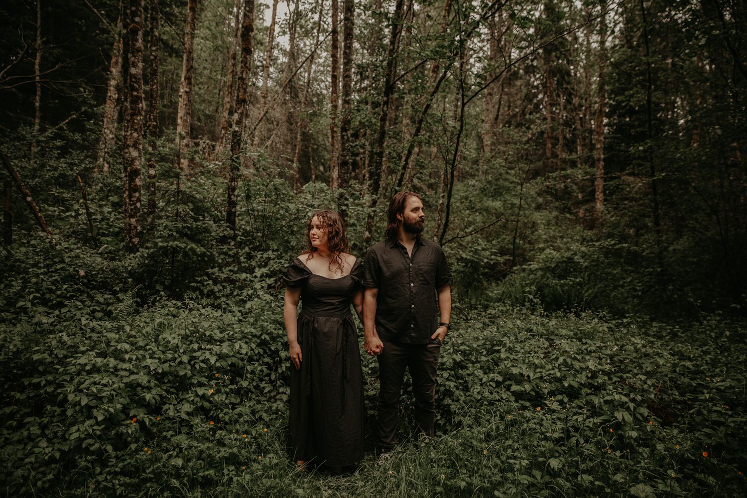 A couple walks through a lush forest path surrounded by dense green foliage and tall trees.