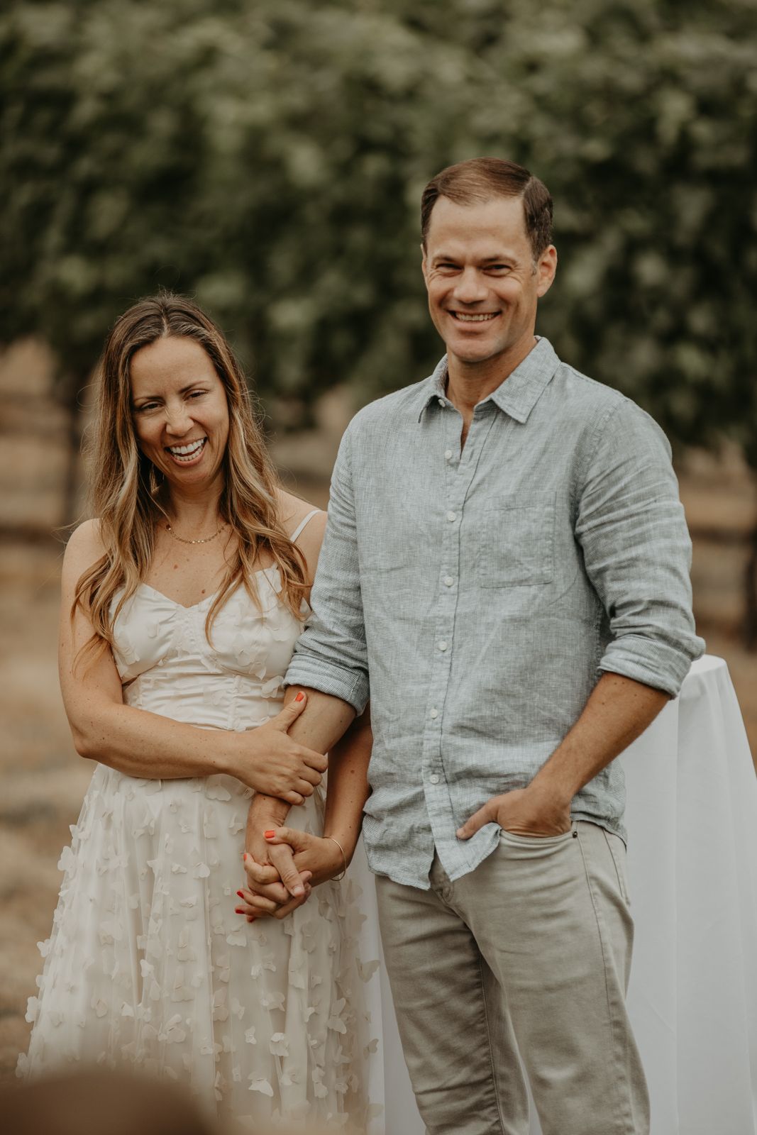 A couple in light-colored outfits shares candid laughter and embraces during an outdoor photo session.