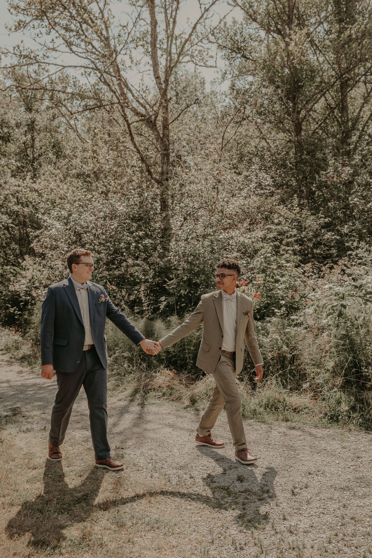 A romantic couple in suits holding hands on a sunlit trail surrounded by blooming spring trees.