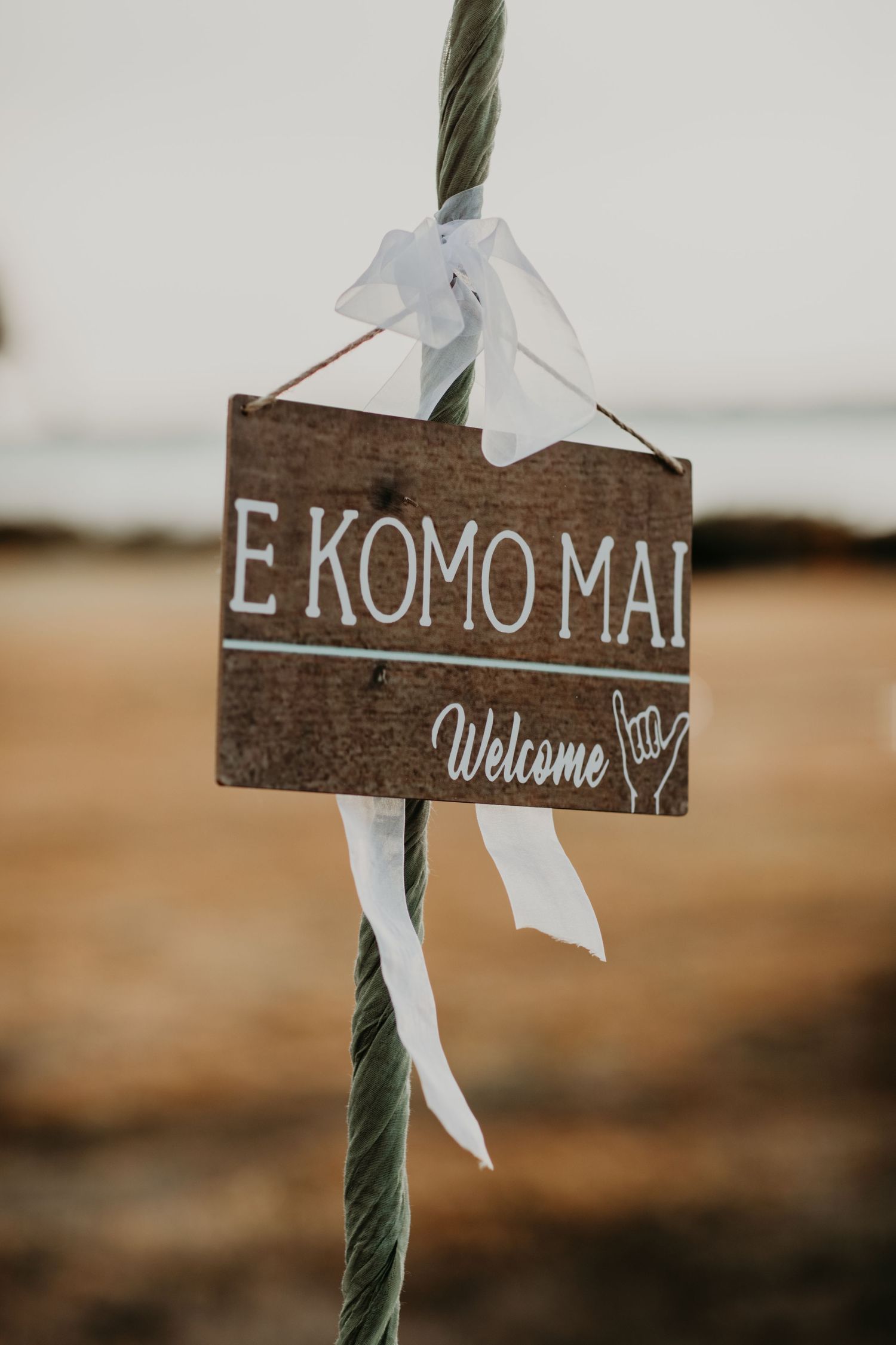 A wooden welcome sign with 'E KOMO MAI' written on it and white ribbons tied to a rope, captured in warm lighting.