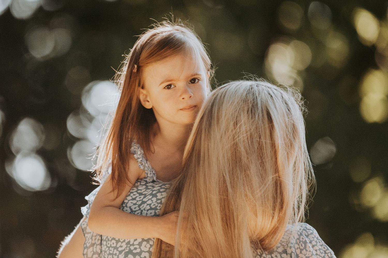 Child with long red hair in silver dress being held up against sunlit bokeh background.